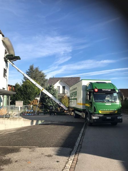 Umzugswagen mit Hebebühne, der bis zu einem Gebäude reicht; grüner LKW auf der Straße, blauer Himmel.
