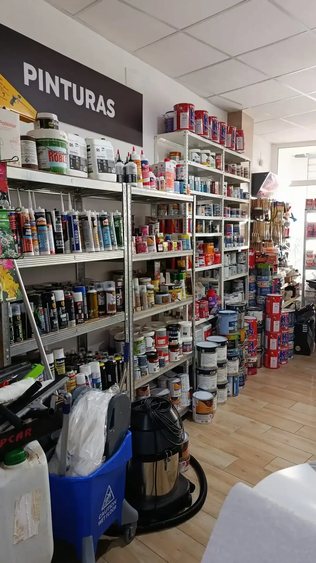 Vista interior de una ferretería con estantes repletos de latas de pintura y suministros, y un cartel de 