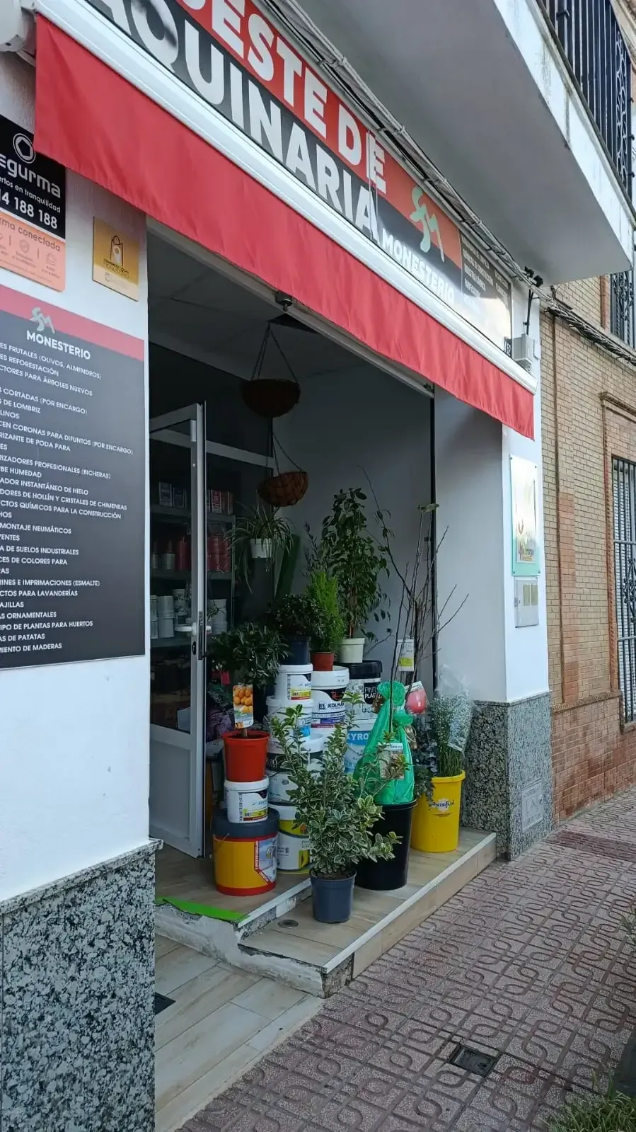 Entrada de una tienda con un toldo rojo. Varias plantas y cubos de pintura expuestos en la puerta.