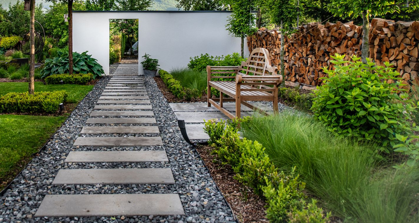 Un chemin de pierres mène à un mur blanc percé d'une ouverture. Un banc en bois et de la verdure bordent le chemin.