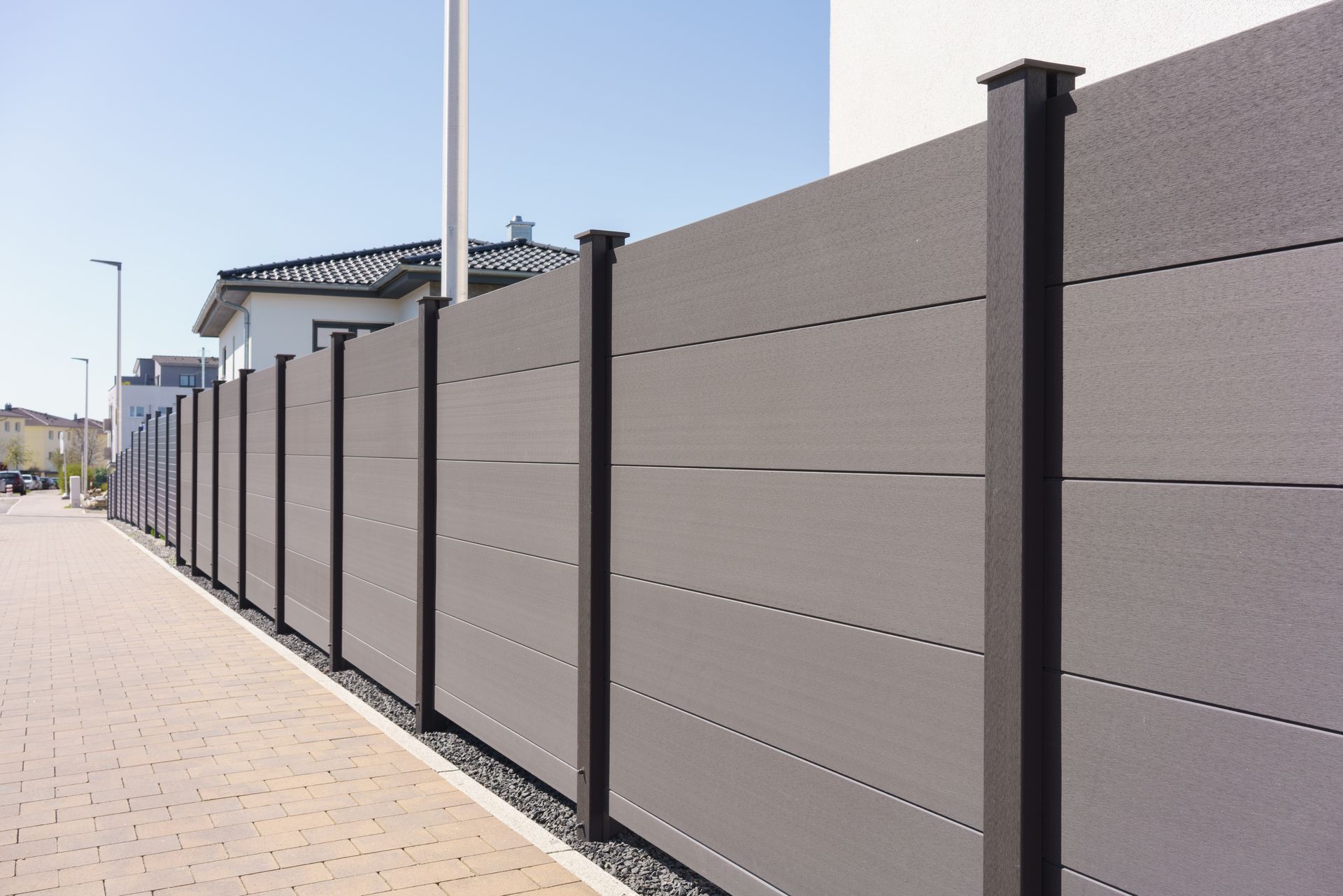Clôture grise à lames horizontales le long d'un trottoir, avec des maisons en arrière-plan par une journée ensoleillée.