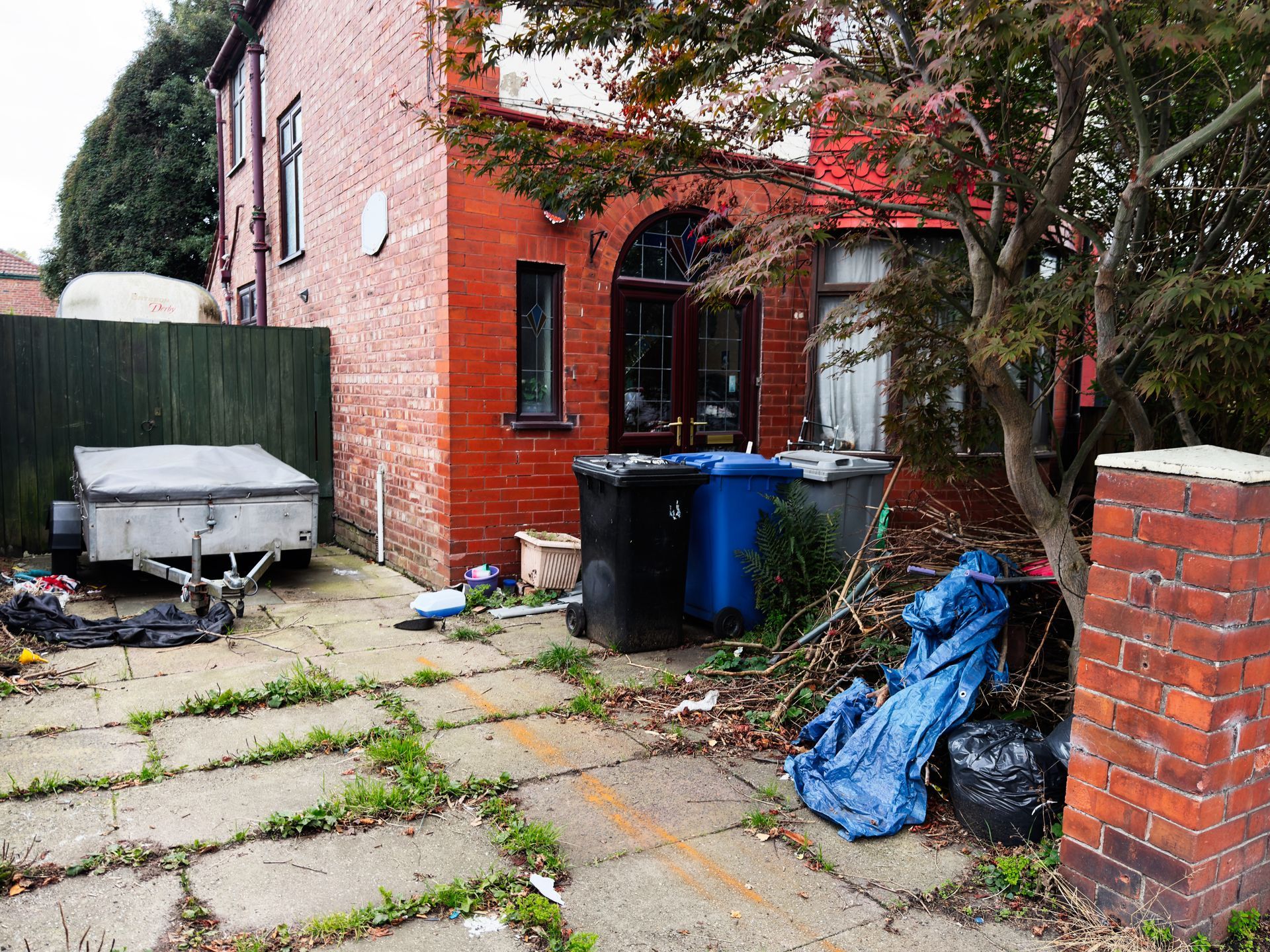 Extérieur d'un bâtiment en briques avec des poubelles débordantes, des débris végétaux sur une zone pavée et une remorque.