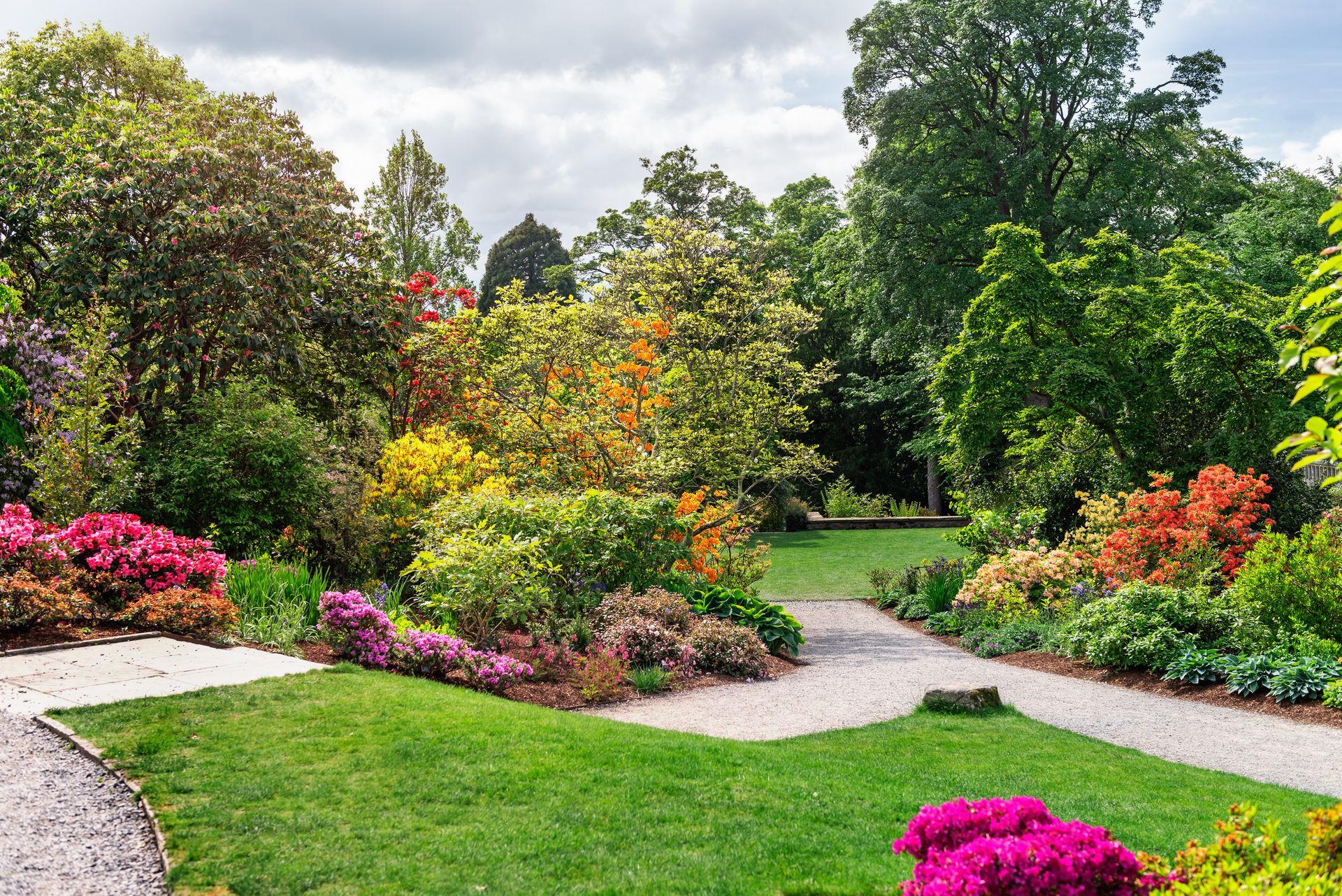 Un jardin avec des allées, une végétation luxuriante, des fleurs roses et orange et des arbres sous un ciel nuageux.