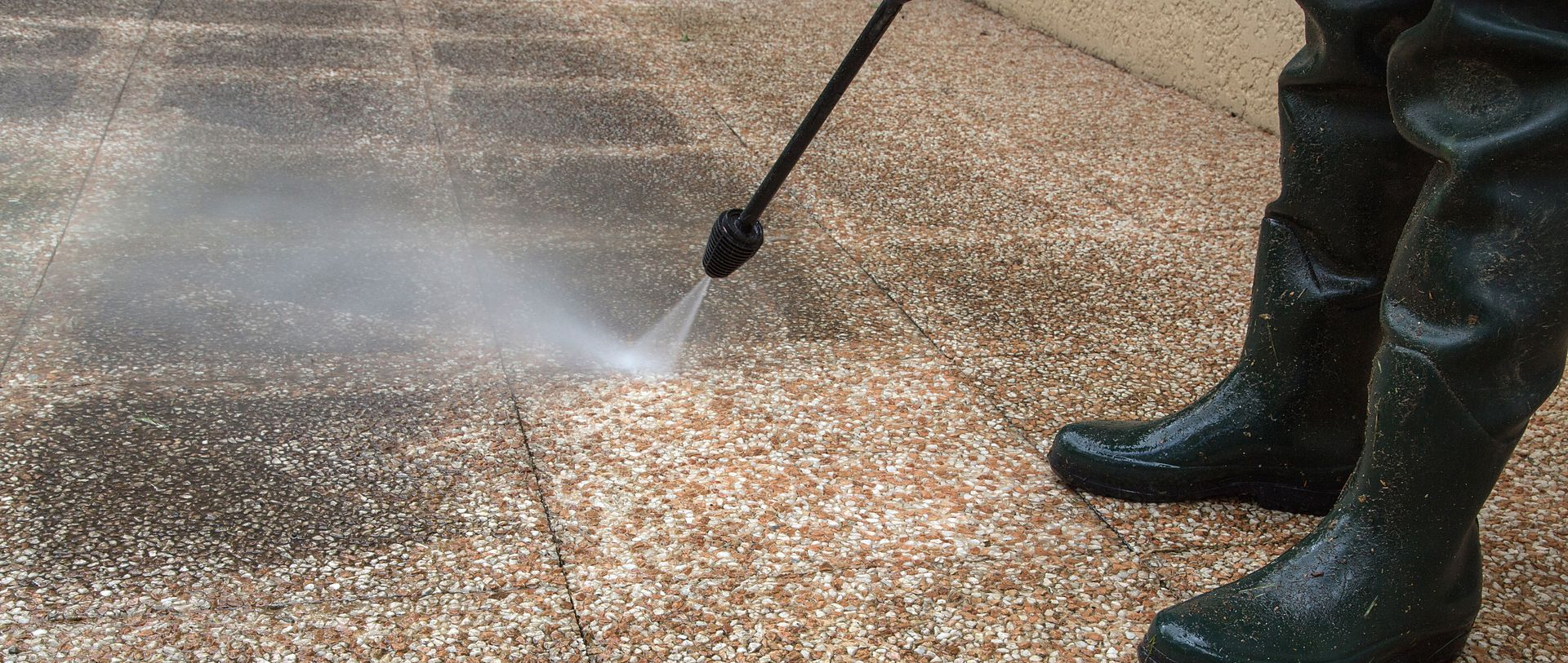 Personne nettoyant au nettoyeur à haute pression une terrasse en pierre, portant de hautes bottes imperméables.