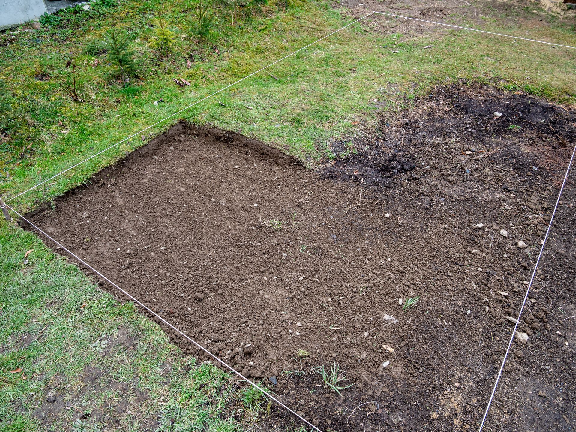 Un parterre de jardin préparé, de forme carrée, avec de la terre foncée et des repères de ficelle blanche sur l'herbe.
