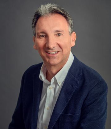 Un hombre con una chaqueta azul y camisa blanca sonríe a la cámara.