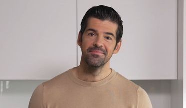 Un hombre con barba está de pie en una cocina vistiendo un suéter color canela.