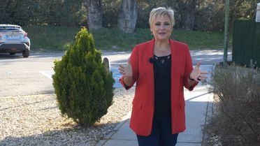 Una mujer con una chaqueta roja está parada en una acera junto a un arbusto.