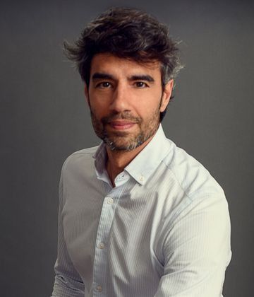 Un hombre con barba lleva una camisa blanca y mira a la cámara.