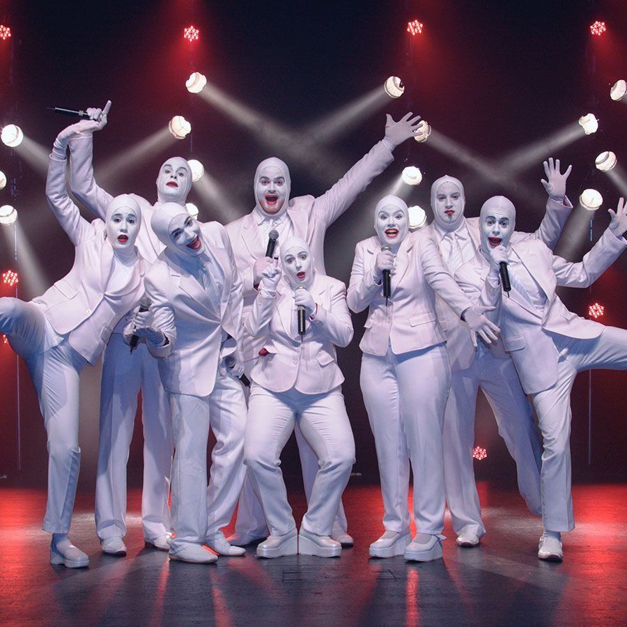 Un grupo de personas vestidas con trajes blancos y máscaras cantan con micrófonos.
