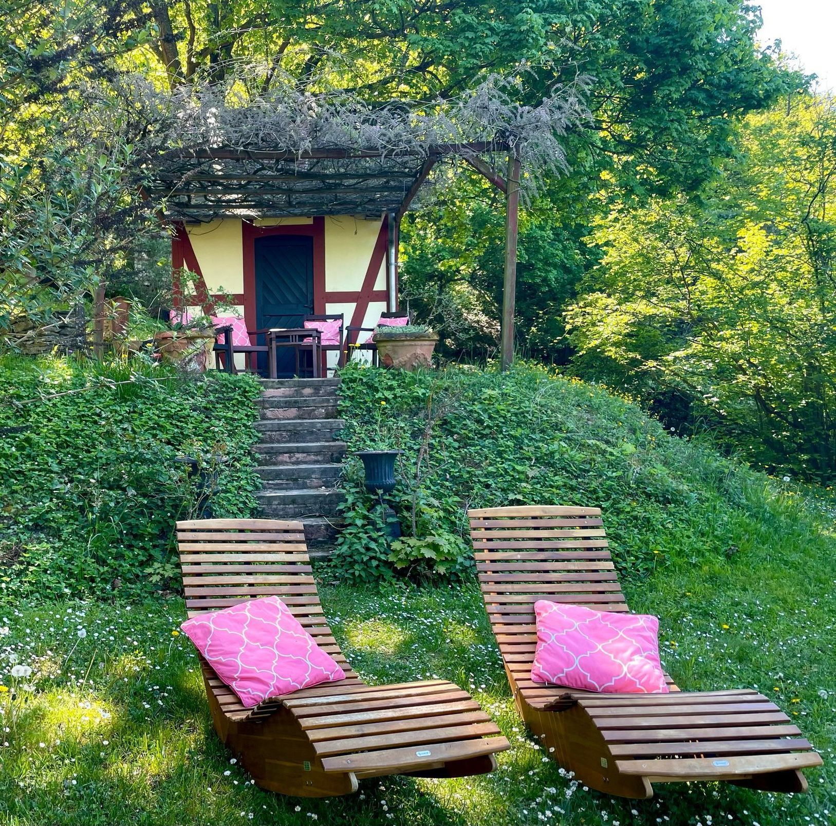Zwei hölzerne Loungesessel mit rosa Kissen vor einem kleinen Haus