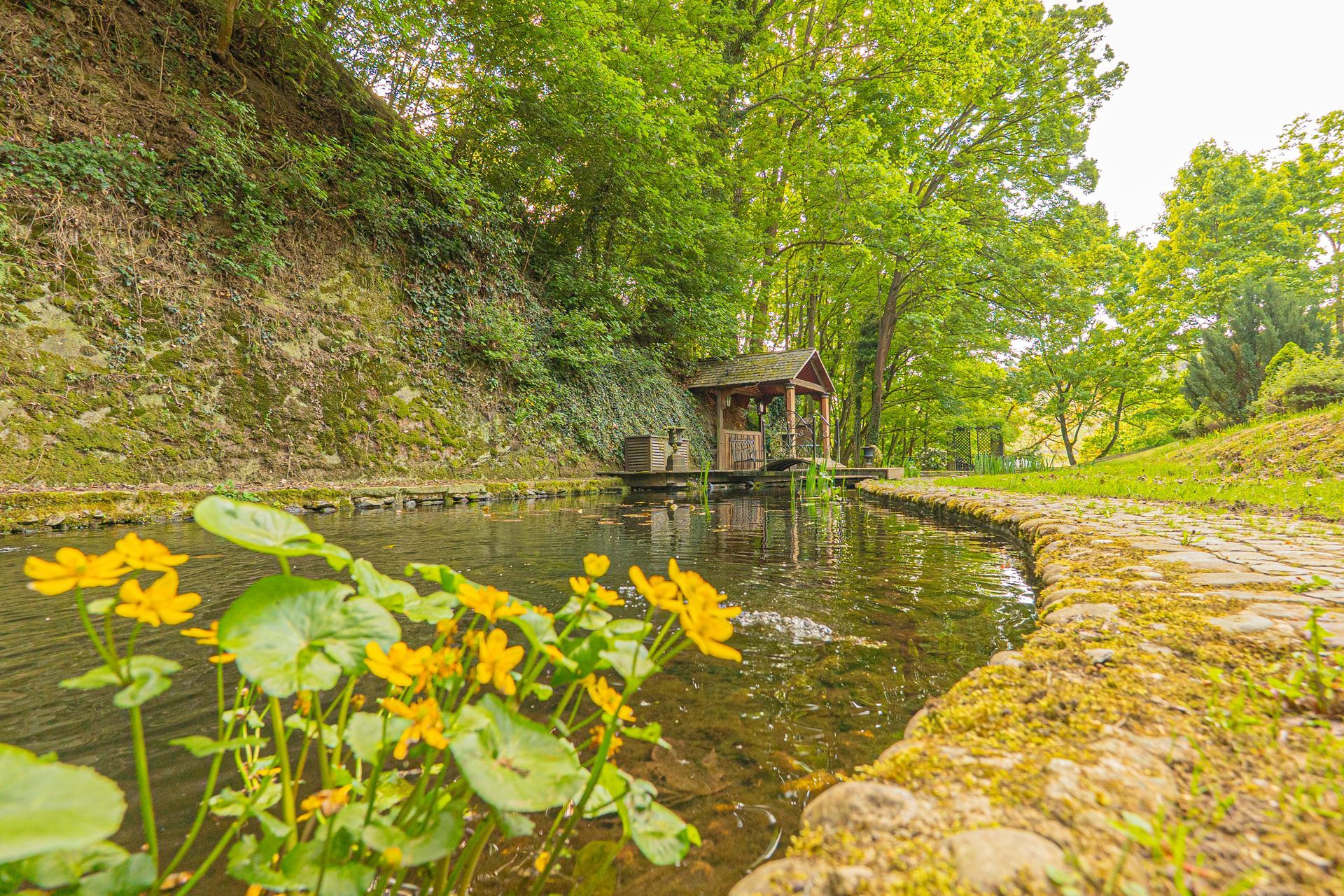 Ein von Bäumen und Blumen umgebener Teich mit einem Pavillon im Hintergrund.