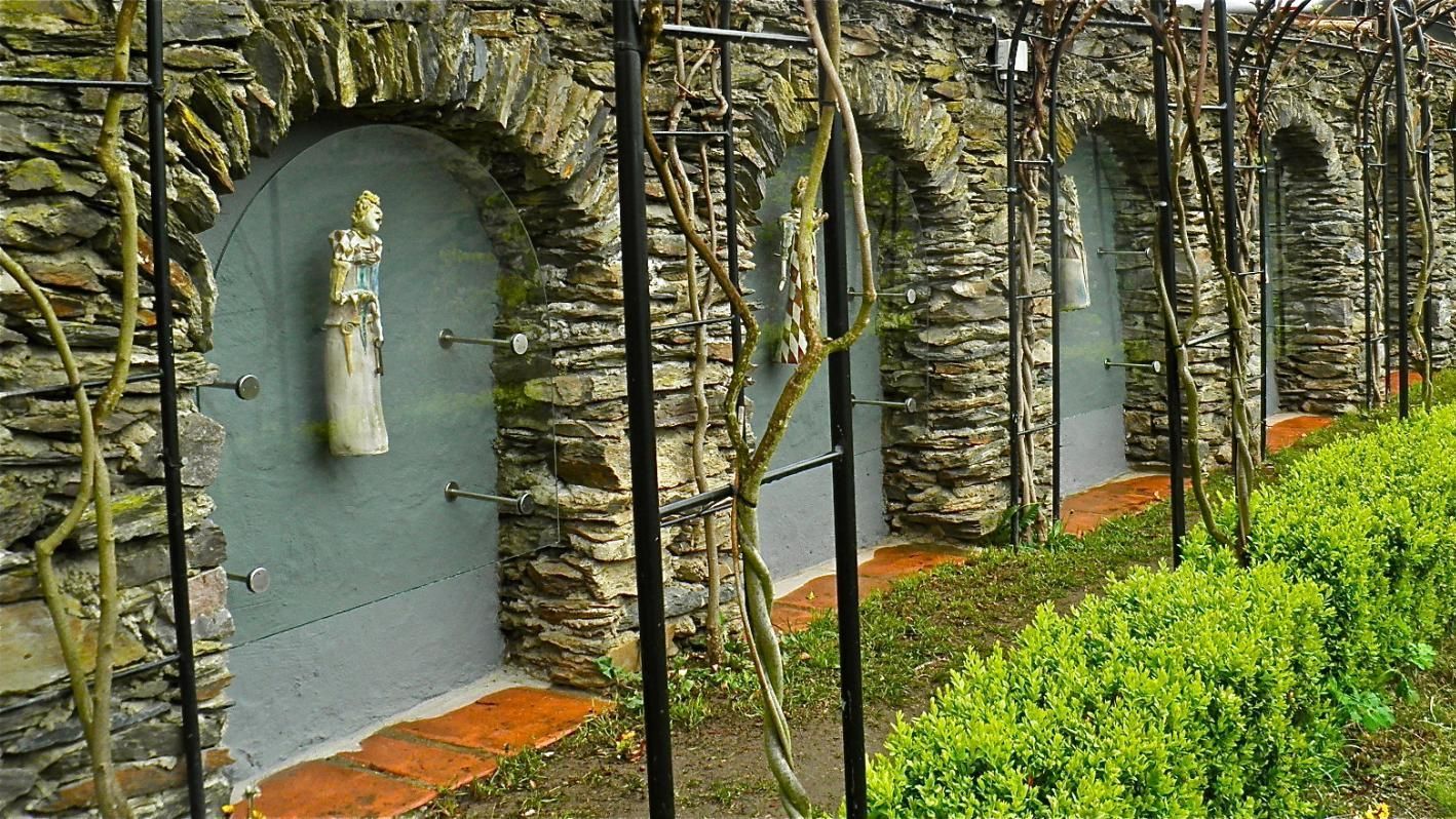 Eine Steinmauer mit Bögen und Büschen mit Skulpturen der sieben Jungfrauen.