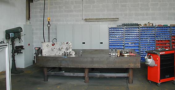 Intérieur d'atelier avec un établi supportant des pièces de machines. Des armoires et des étagères sont visibles en arrière-plan.