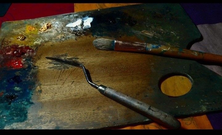 Dos pinceles sobre una paleta de artista salpicada de pintura de colores mezclados y un agujero en un vaso de pintura amarilla.
