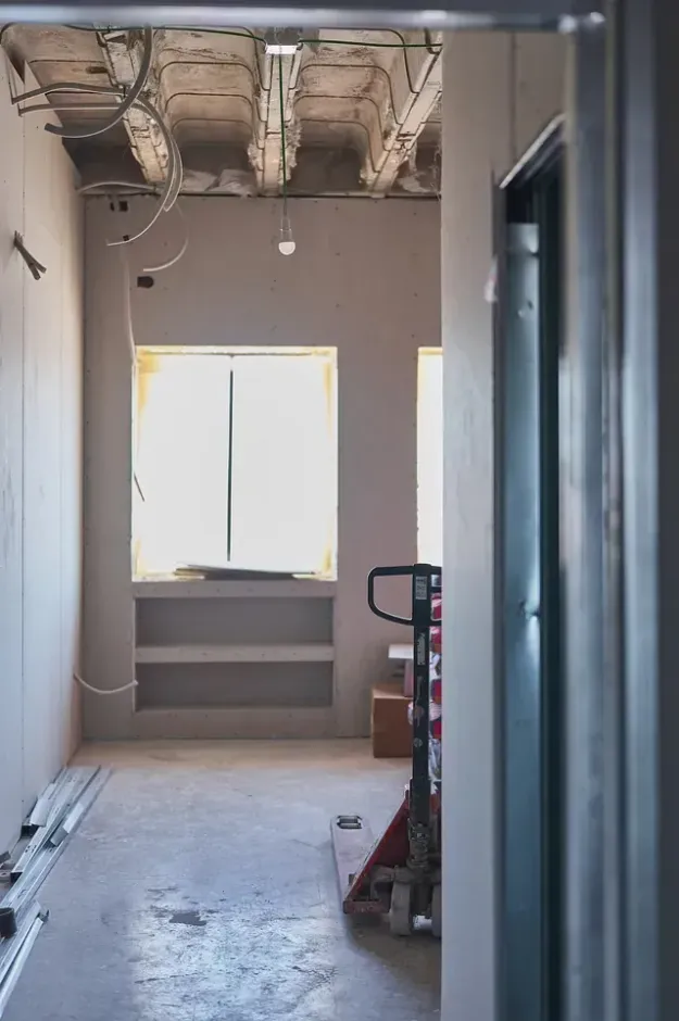Construcción de un pasillo estrecho con ventana, estanterías y transpaleta. Paredes grises y cableado del techo a la vista.