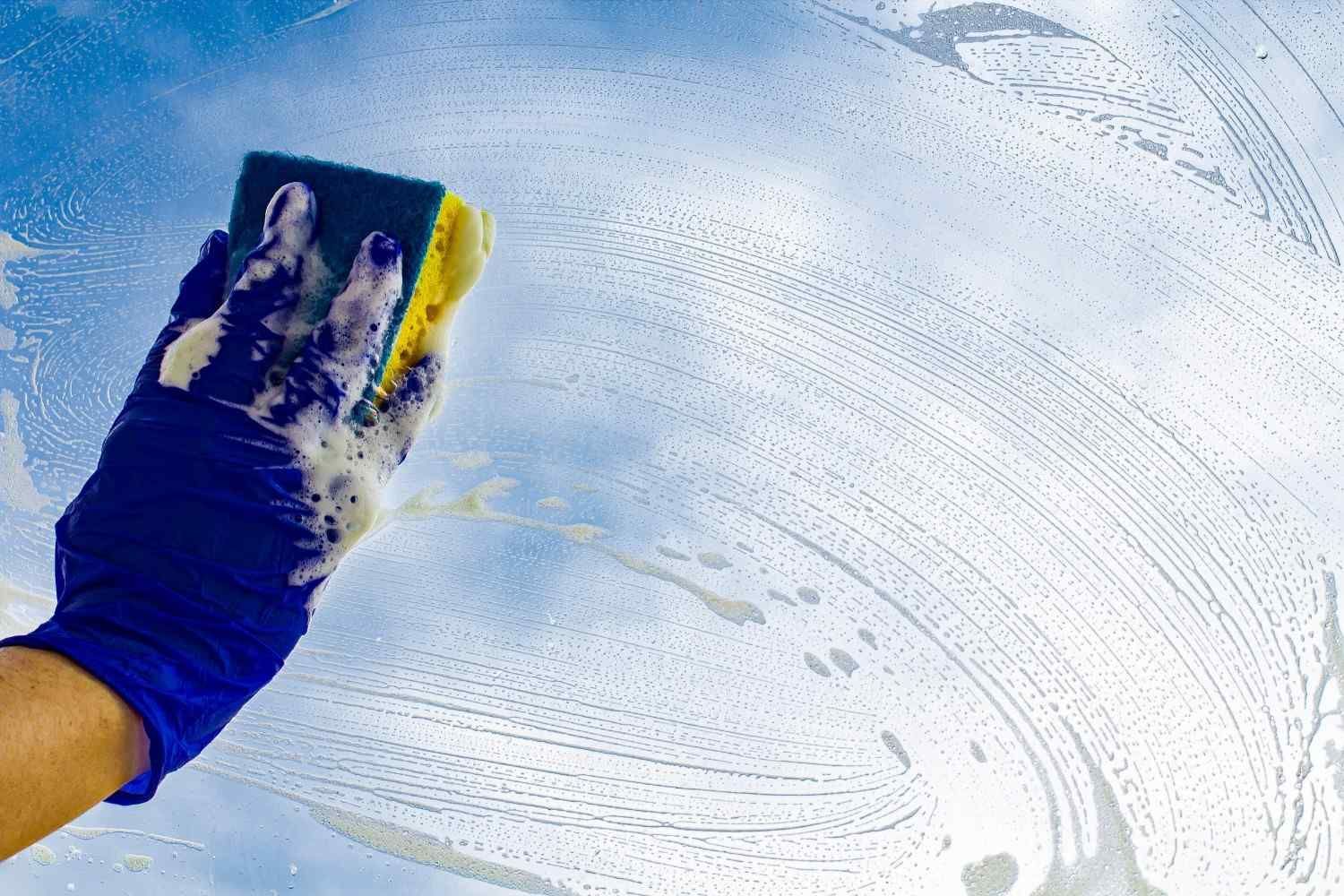 Una mano enguantada limpia una ventana con un limpiador jabonoso, dejando manchas blancas en el cristal.