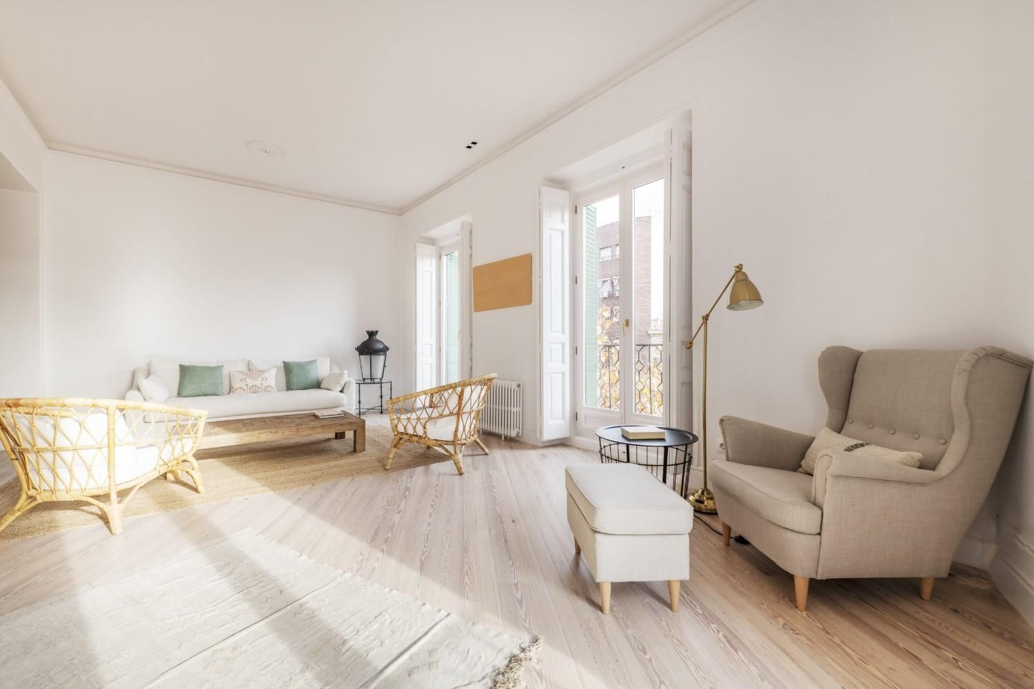 Luminoso salón minimalista con sofá color crema, sillas de mimbre, lámpara de pie y luz natural que entra por los grandes ventanales.