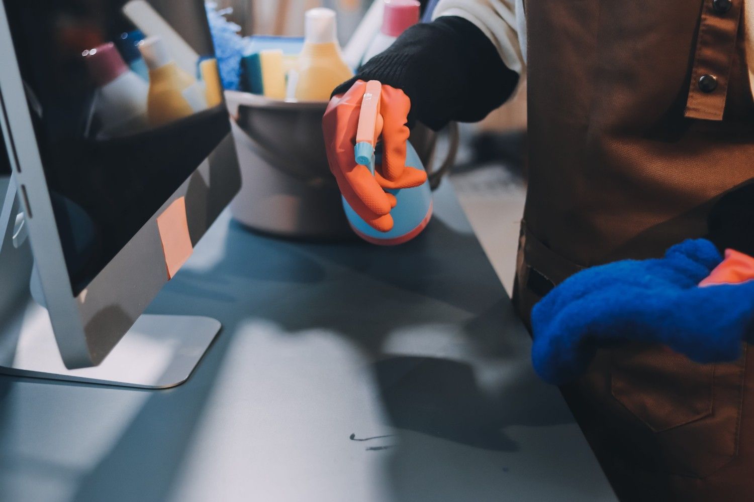 Manos enguantadas limpiando una estación de trabajo informática con un pulverizador y un paño.