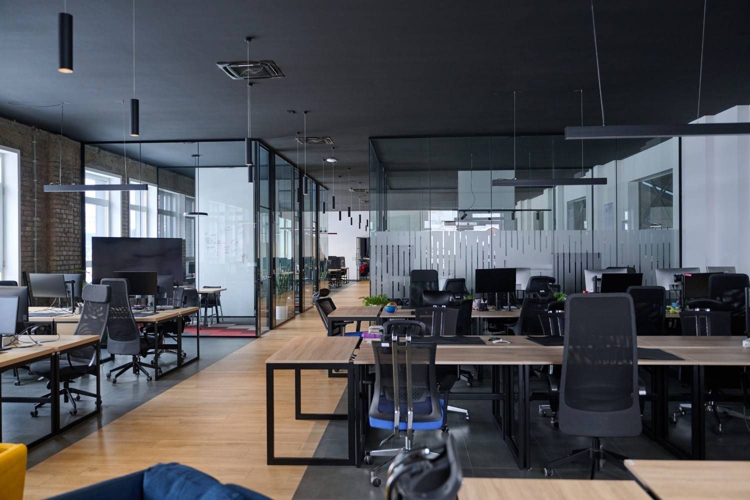 Oficina moderna de planta abierta con escritorios, sillas, ordenadores y paredes de cristal, visible desde un pasillo central.