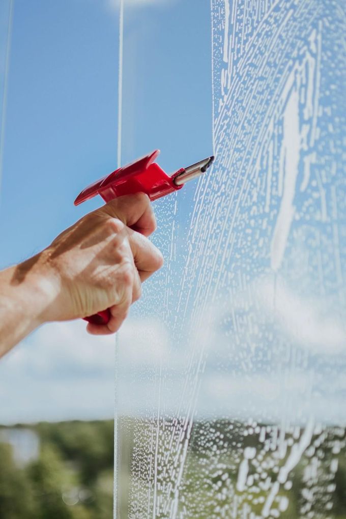 Una mano usa una espátula roja para limpiar una ventana de vidrio con gotas de agua.