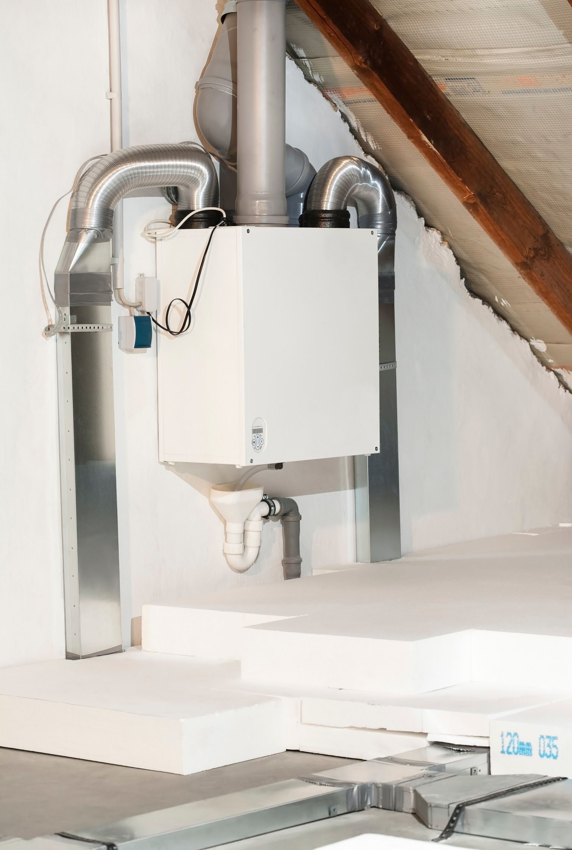 Un système de ventilation HRV monté sur un mur de grenier avec des conduits métalliques, relié à des panneaux d'isolation de sol blancs situés en dessous.
