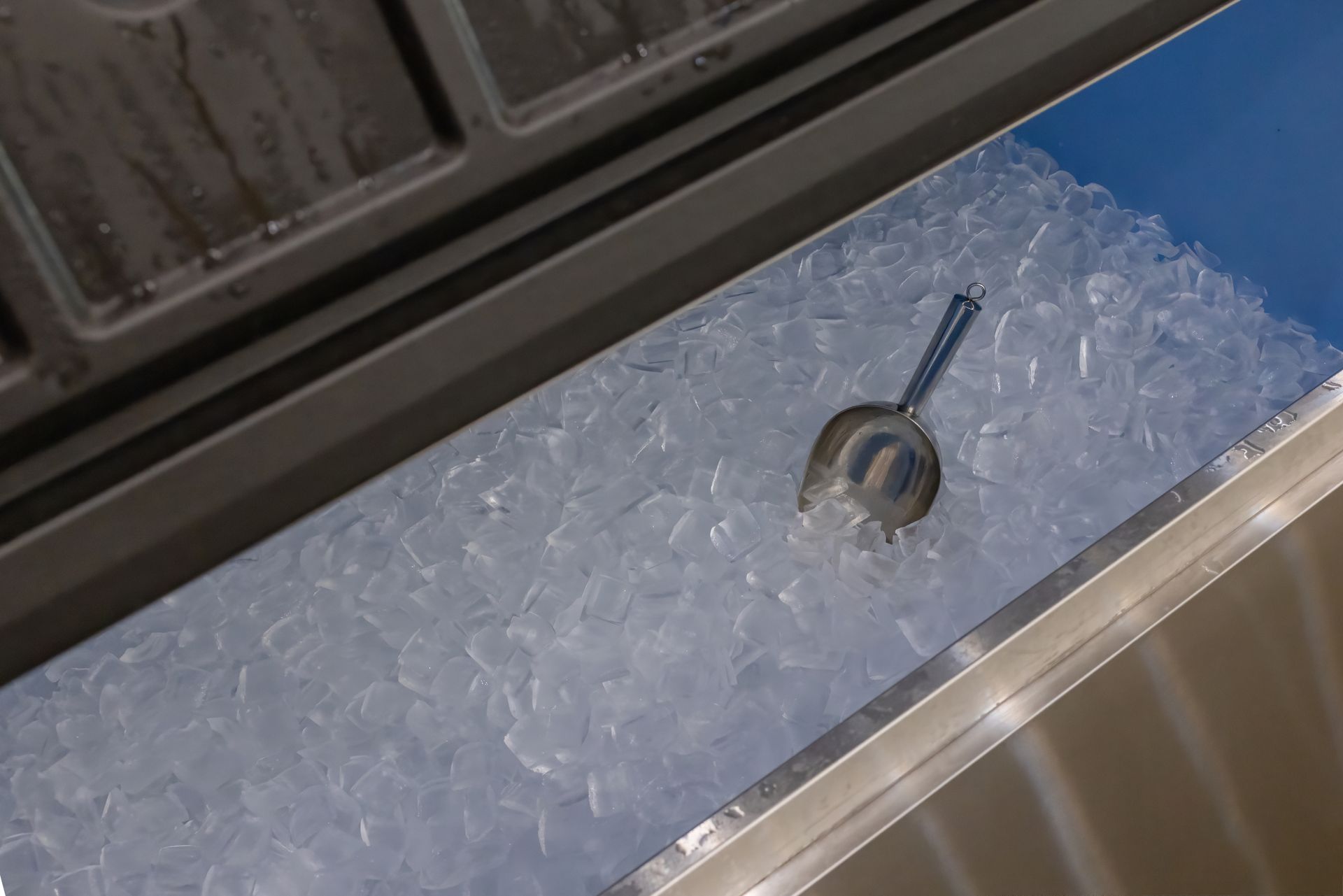 Une pelle à glace en métal repose dans un grand bac rempli de glaçons transparents.