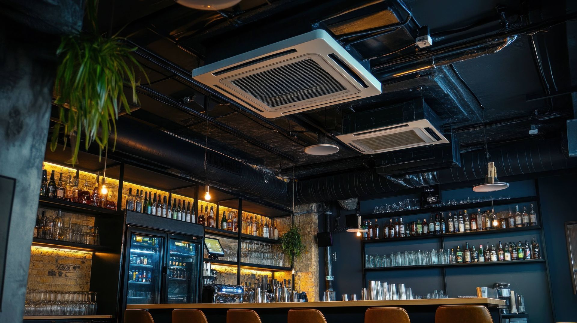 Un bar moderne à l'éclairage tamisé, avec des plafonds sombres, deux bouches d'aération carrées, des étagères remplies de bouteilles et un comptoir en bois.