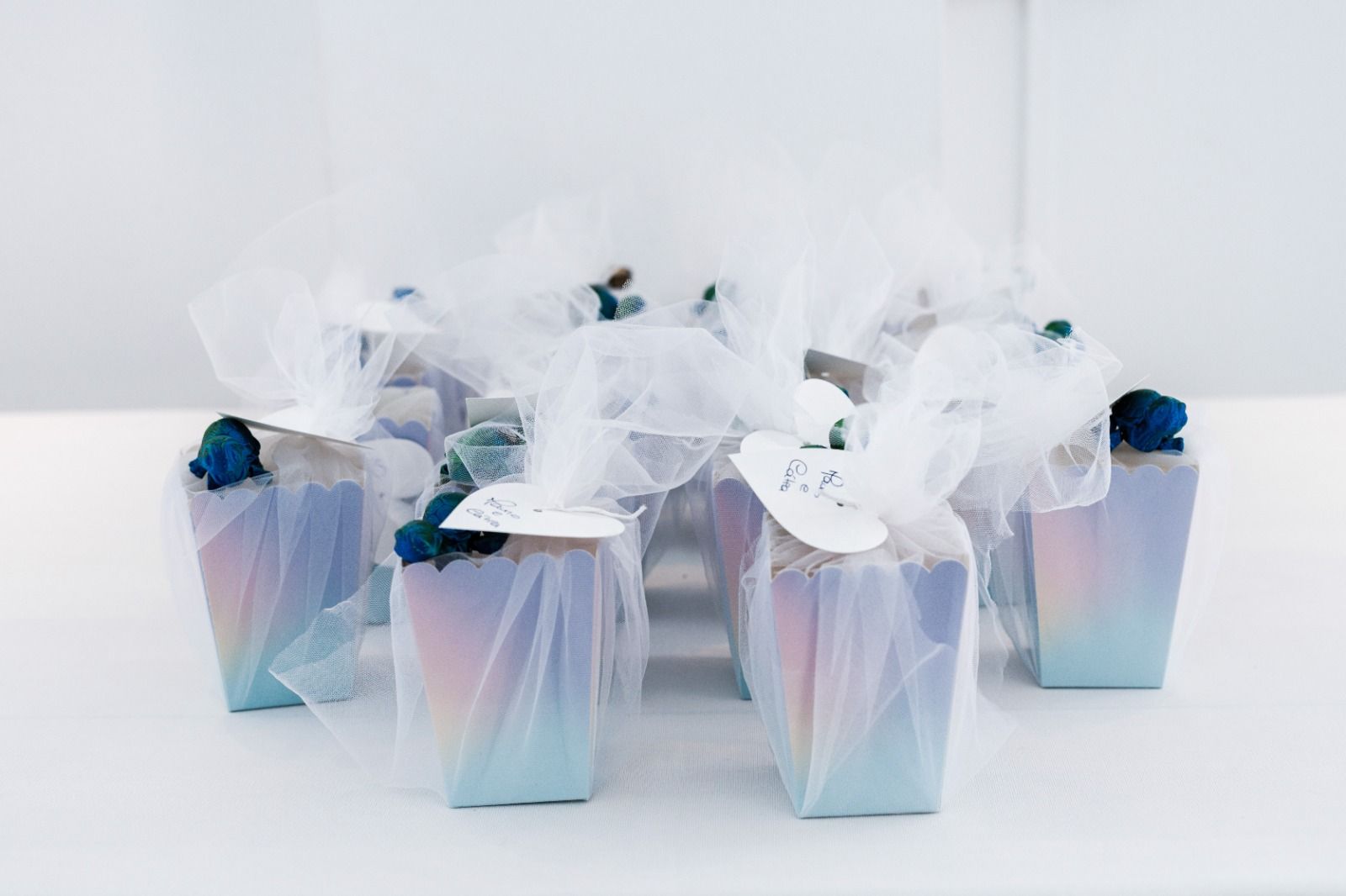 Pequeñas y coloridas cajas de regalo envueltas en tul blanco y adornadas con una pequeña flor azul.