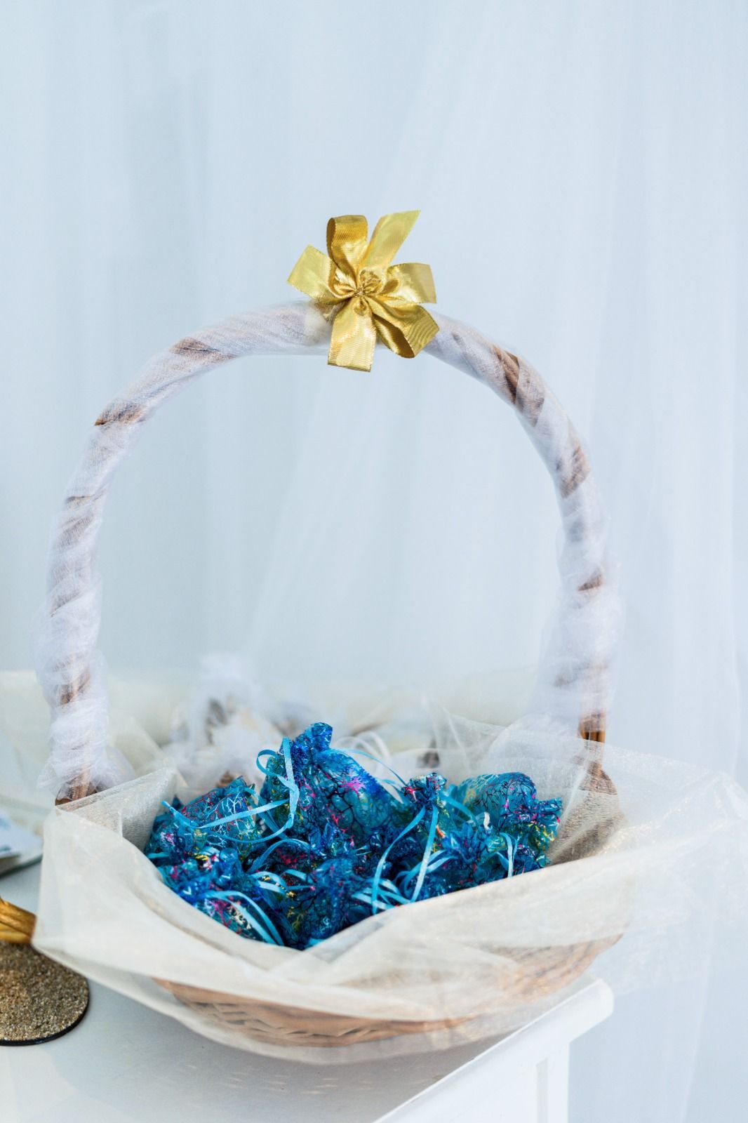 Cesta de mimbre con un lazo dorado en la parte superior, llena de caramelos azules, colocada sobre una superficie blanca.