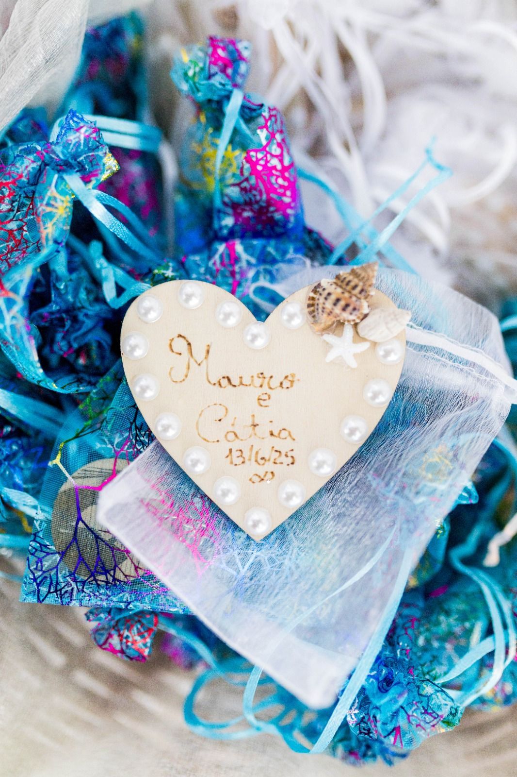 Etiqueta en forma de corazón con nombres y fecha, conchas marinas, cintas azules y tela en una cesta.