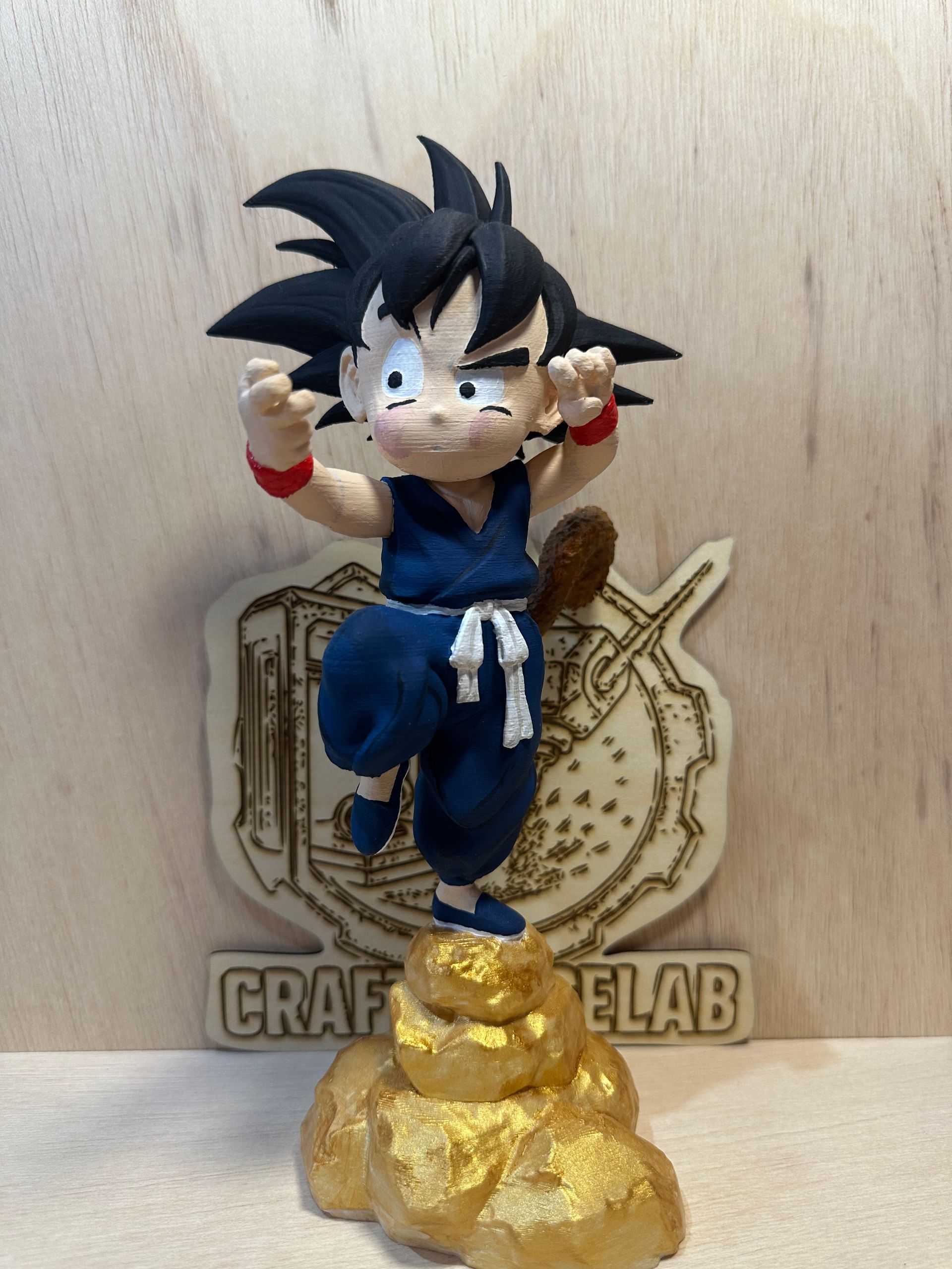 Figura de Goku niño con gi azul, base de nube dorada, sobre un fondo de madera con un logotipo.