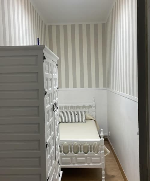 Dormitorio estrecho con muebles blancos, pared decorativa a rayas y suelo de madera.