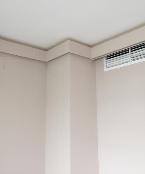 Esquina de una habitación con paredes color beige, techo blanco y moldura cuadrada. Hay una rejilla de ventilación en la esquina.