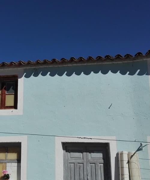 Edificio azul con ventana y puerta, tejas oscuras contra un cielo azul claro.