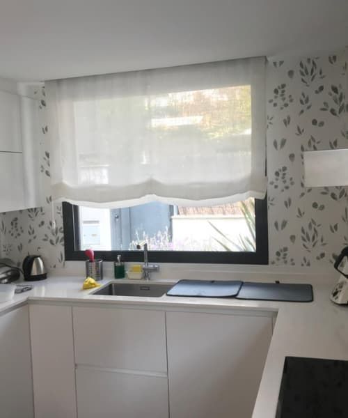 Cocina con armarios blancos, fregadero y ventana con una cortina blanca traslúcida. El papel pintado tiene un sutil estampado de hojas.