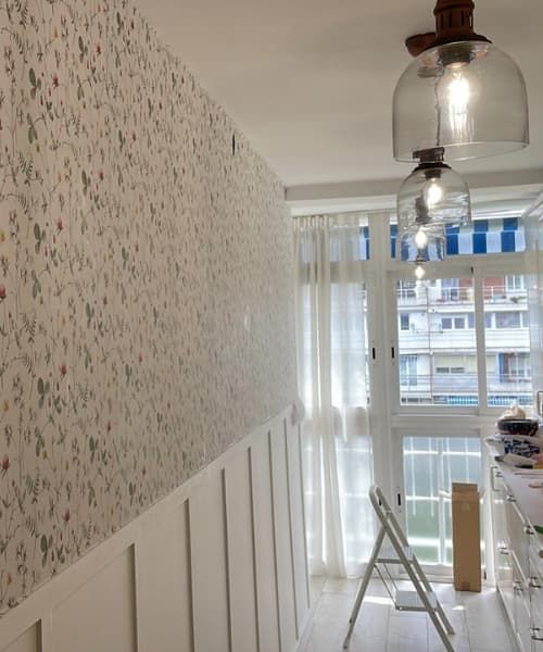 Una habitación con papel pintado floral y paneles de madera blancos. Dos lámparas colgantes de cristal iluminan una ventana con una cortina transparente.