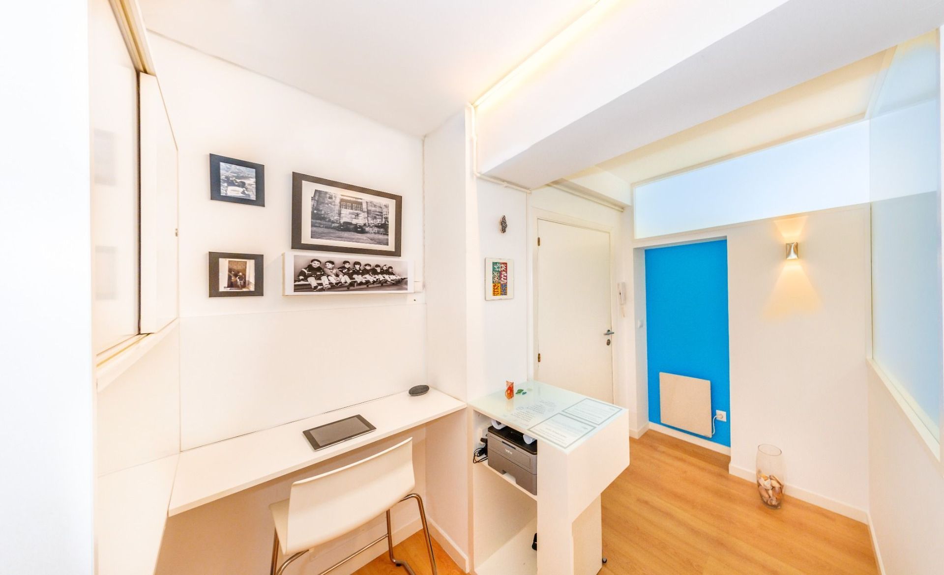 Pasillo blanco luminoso con fotos enmarcadas, un pequeño escritorio, escalera y pared decorativa azul en una casa moderna.
