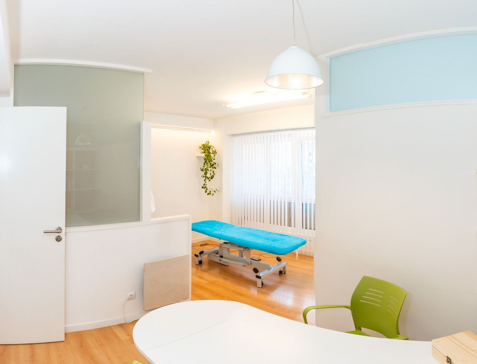 Sala de exploración médica luminosa con camilla de tratamiento, paredes blancas y silla verde cerca de una mesa redonda.