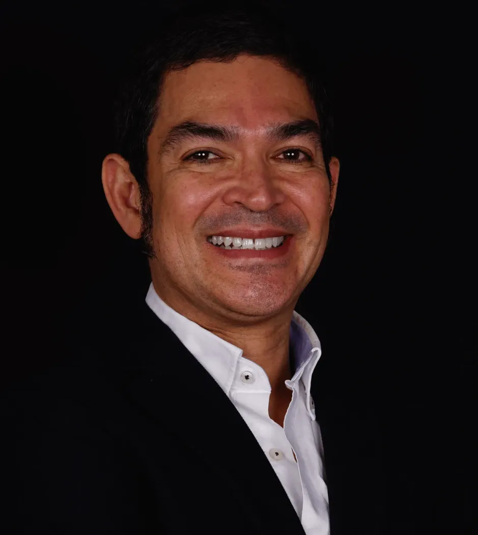 Hombre sonriente con traje negro y camisa blanca sobre fondo oscuro.