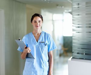 Enfermera con uniforme azul, sosteniendo un portapapeles, sonriendo en un pasillo del hospital.