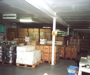 Interior de almacén con cajas apiladas sobre paletas, una columna de soporte e iluminación cenital.