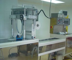 Máquina industrial con estructura y controles metálicos, que dispensa material sobre una cinta transportadora en una sala limpia.