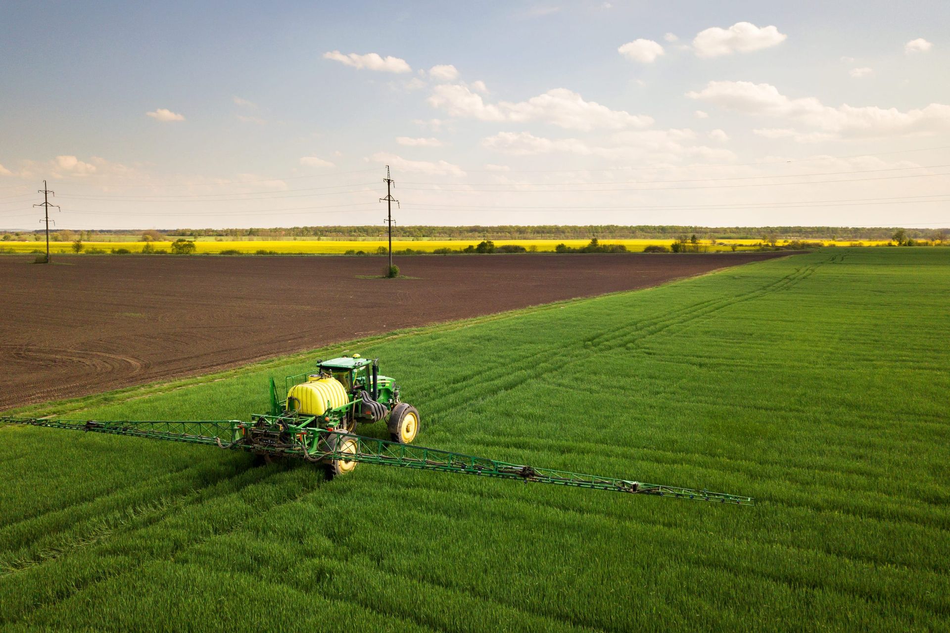 Un tractor verde rocía un campo verde brillante al lado de una parcela oscura y arada bajo un cielo azul con nubes dispersas.