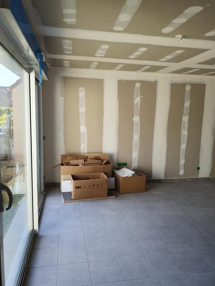 Pièce vide et inachevée avec sol carrelé, plaques de plâtre réparées et cartons près d'une grande fenêtre.