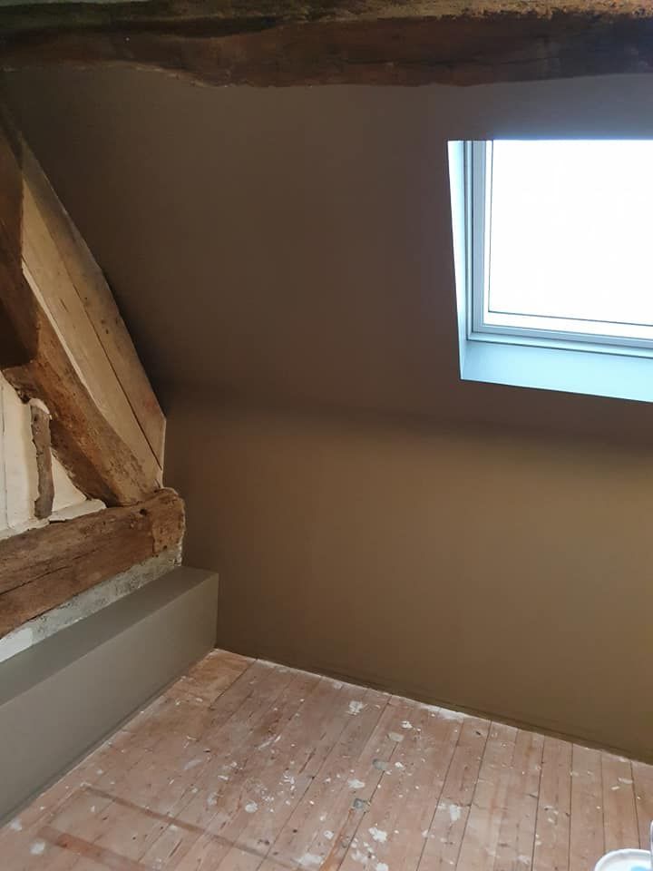 Pièce mansardée avec poutres apparentes, mur brun en pente et une petite fenêtre au-dessus d'un plancher en bois nu.