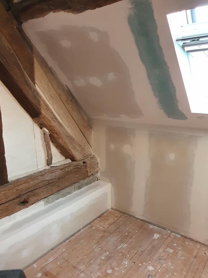 Grenier en rénovation avec poutres apparentes, plaques de plâtre réparées et plafond mansardé près d'une fenêtre.