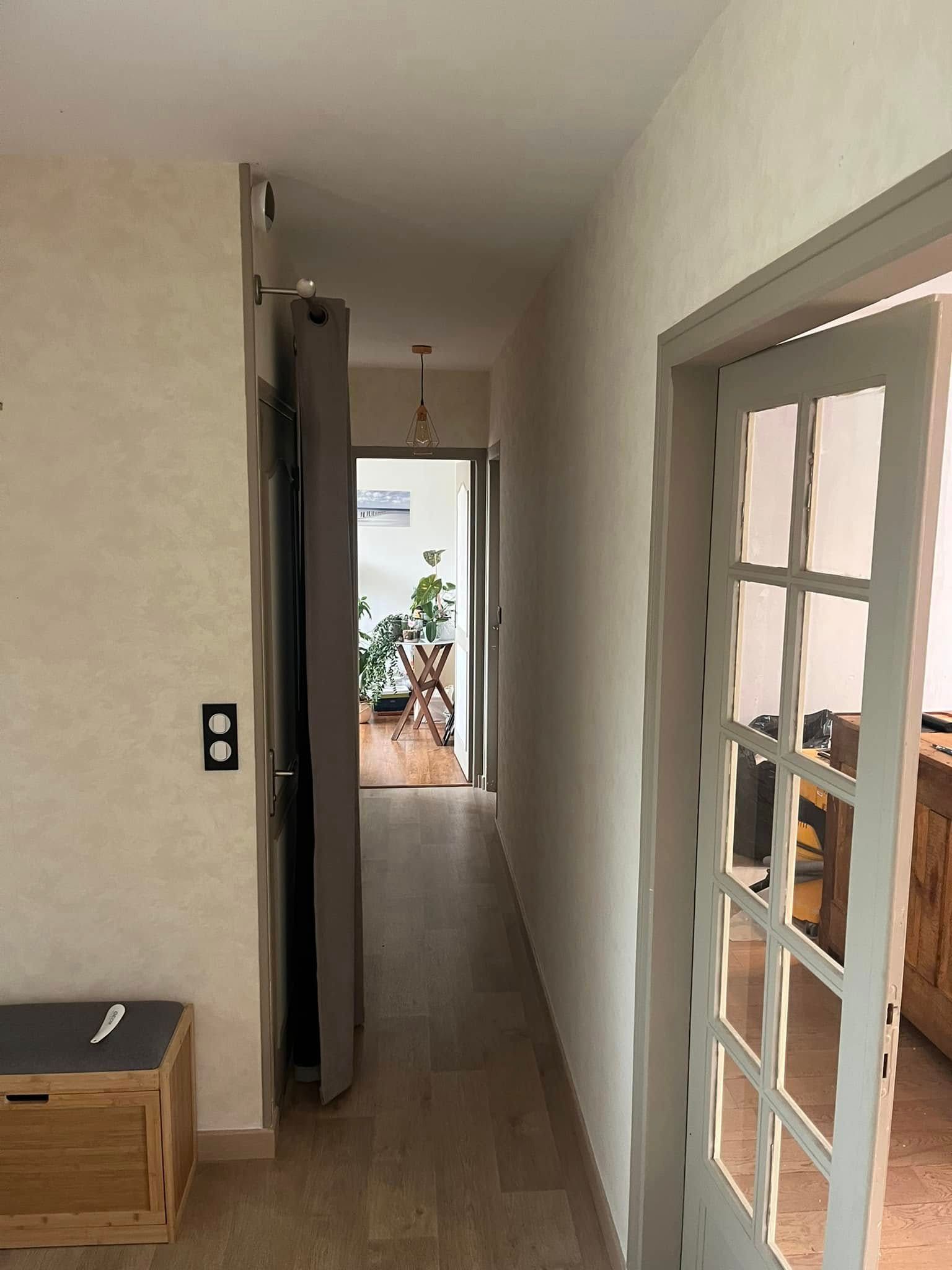 Couloir étroit aux murs clairs, parquet et portes-fenêtres donnant sur un patio lumineux.