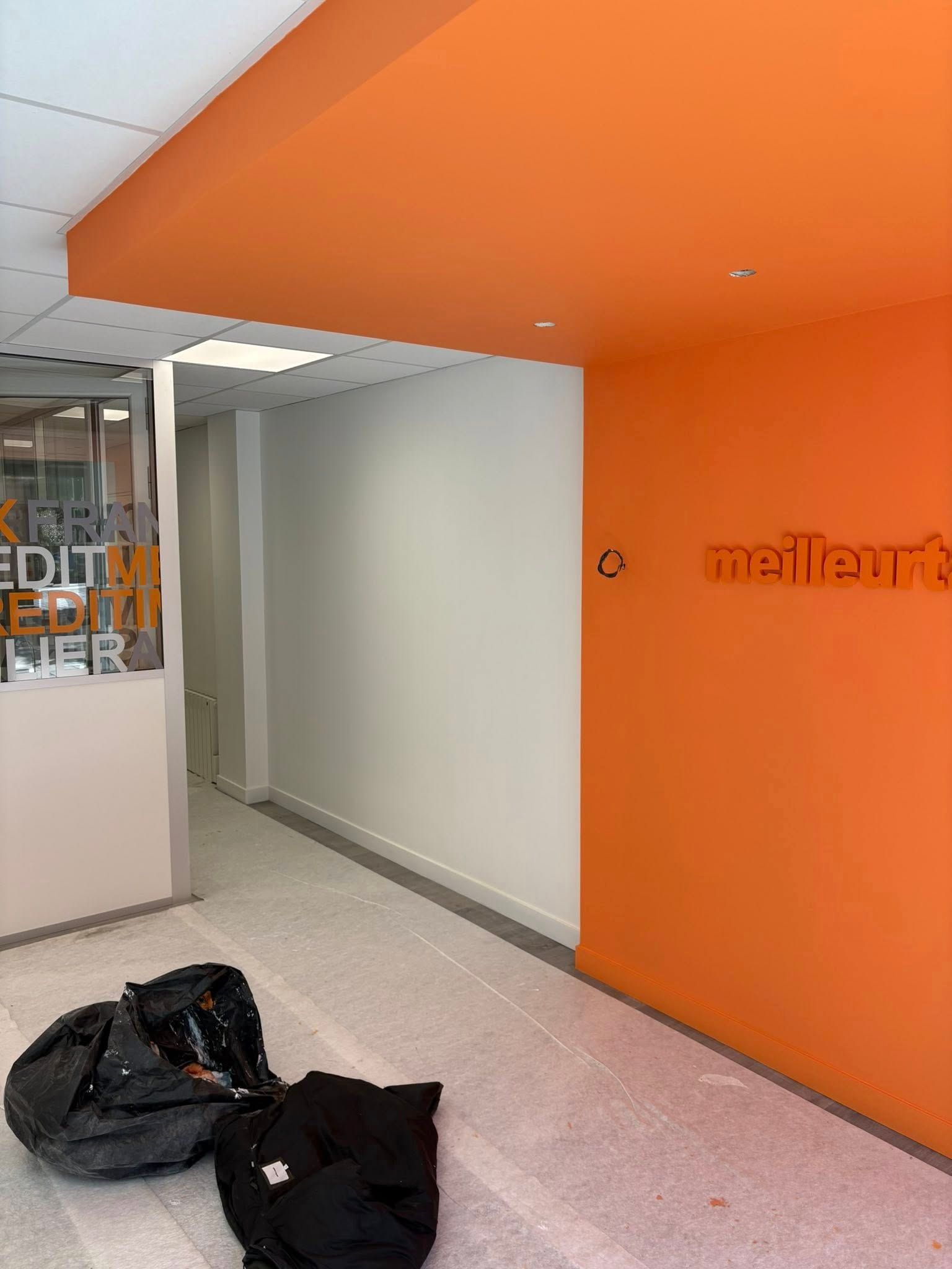Couloir aux murs orange, plafond à dalles apparentes et deux sacs noirs au sol près d'une entrée de bureau vitrée