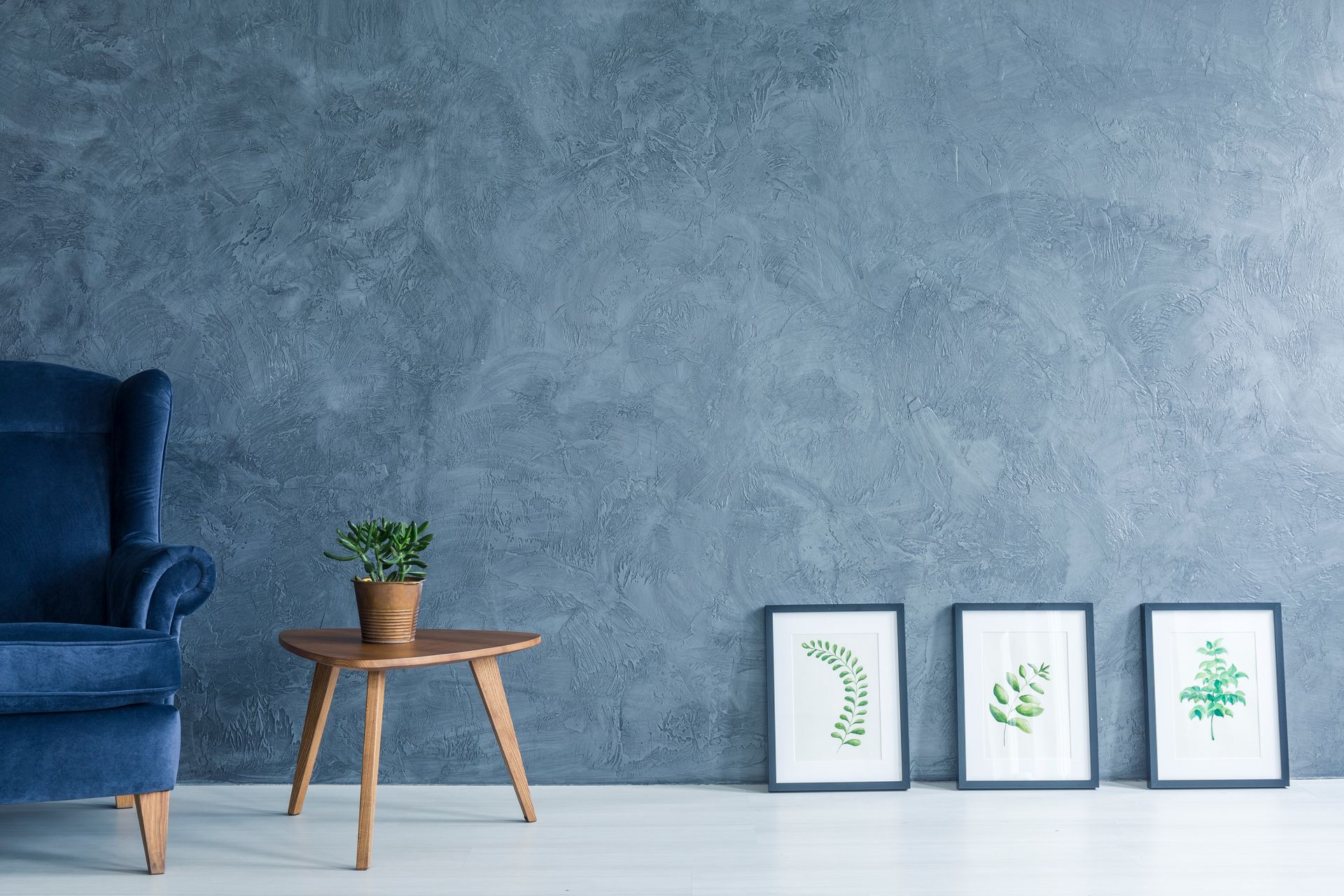 Un canapé bleu à côté d'une table d'appoint en bois et trois estampes de feuilles encadrées sur un mur bleu texturé.